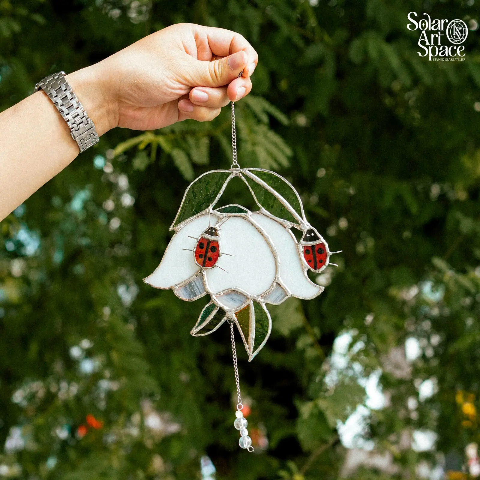 Suncatcher hình bông hoa chuông kính trắng úp xuống, lá xanh phía trên, hai chú bọ rùa đỏ chấm đen đậu trên cánh, chuỗi hạt trong rủ bên dưới, một tay giơ cao trước tán lá