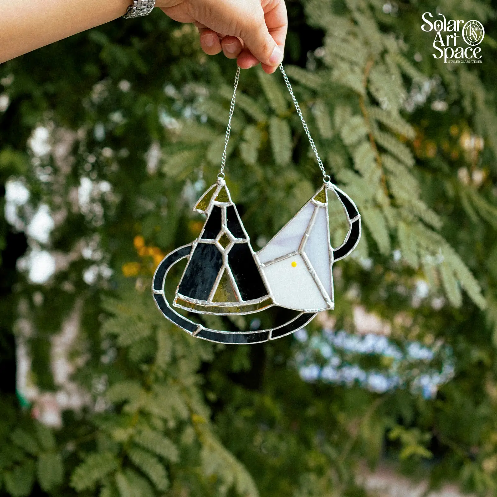Suncatcher gồm một khối kính đen hình nón có núm vàng xanh trên đỉnh và một khối kính trắng có quai đen và một chấm vàng, cả hai đứng trên đế cong bầu dục viền đen, treo dây xích trước tán lá