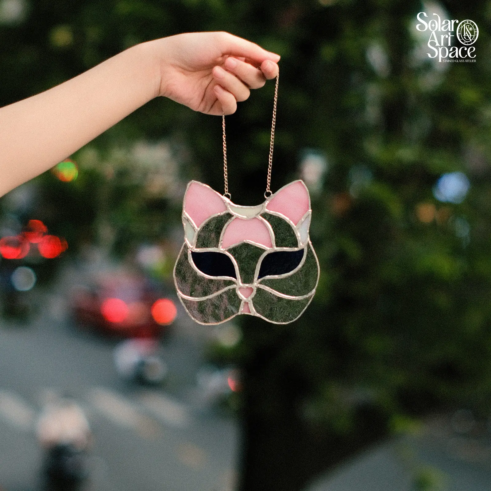 Suncatcher hình mặt mèo nhìn thẳng, má kính xanh rêu vân, tai và vệt trán hồng, hai mắt kính đen xếch, treo dây xích, cầm giơ trước hàng cây