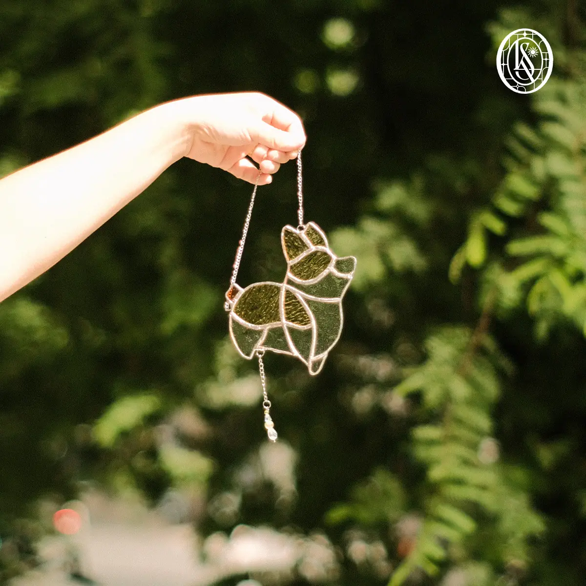 Suncatcher hình chó corgi màu nâu vàng bằng kính vân, bụng trắng, hai tai dựng, một charm nhỏ rủ bên dưới bằng dây, cầm giơ trước tán lá