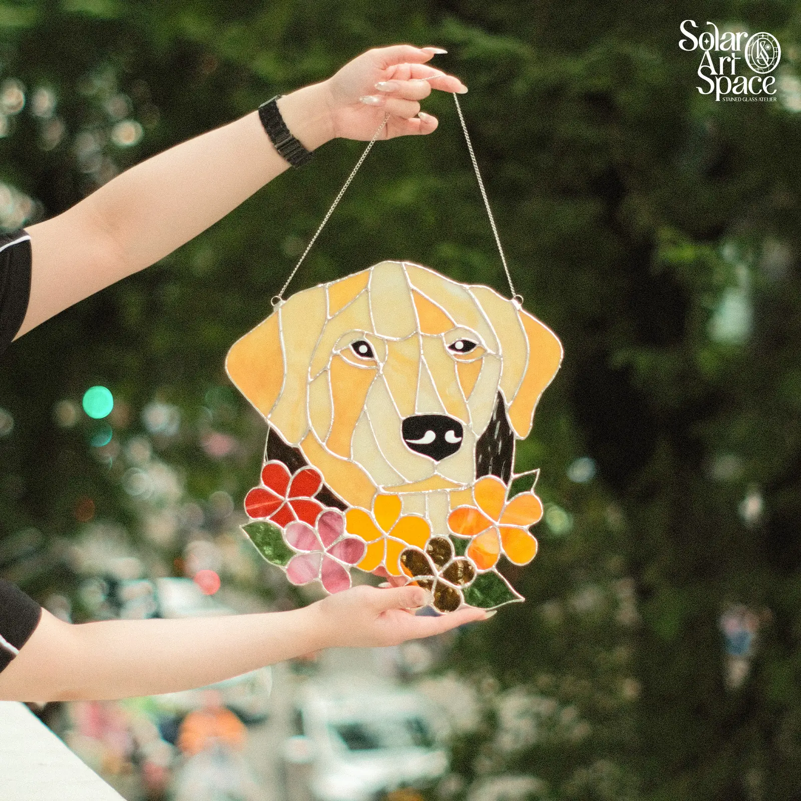 Suncatcher hình đầu chú chó lông vàng nhìn thẳng, tai cam rủ, mũi kính đen, dưới cằm một vòng hoa đỏ, hồng, vàng, cam và nâu hổ phách xen lá xanh, treo dây xích, hai tay cầm trước tán lá