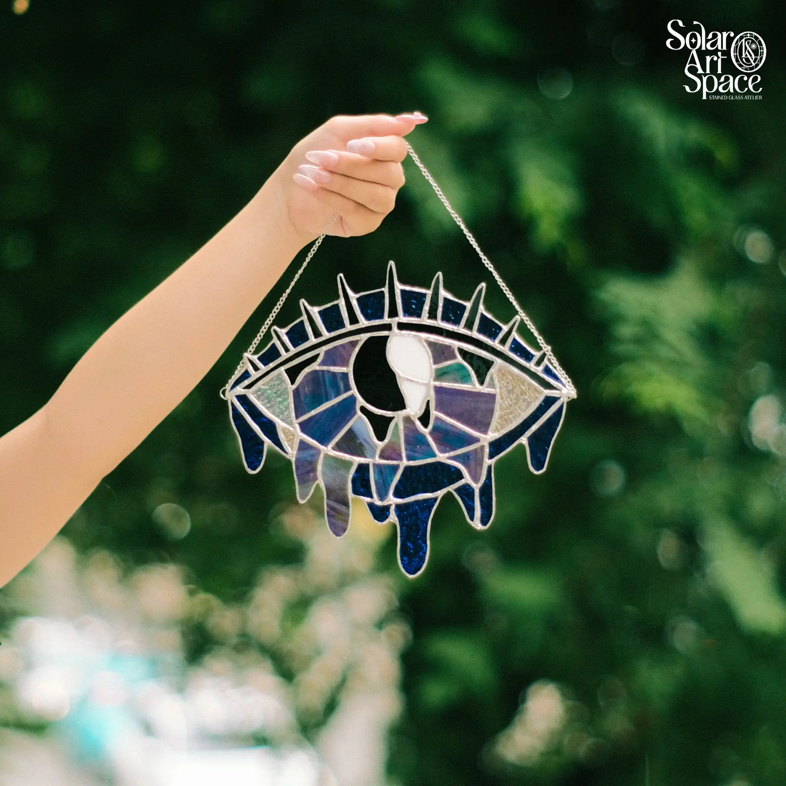 Suncatcher hình con mắt lớn bằng kính xanh tím ánh xà cừ, con ngươi đen trắng, hàng mi nhọn phía trên, những giọt kính xanh đậm chảy xuống mép dưới, treo dây xích, cầm giơ trước tán lá