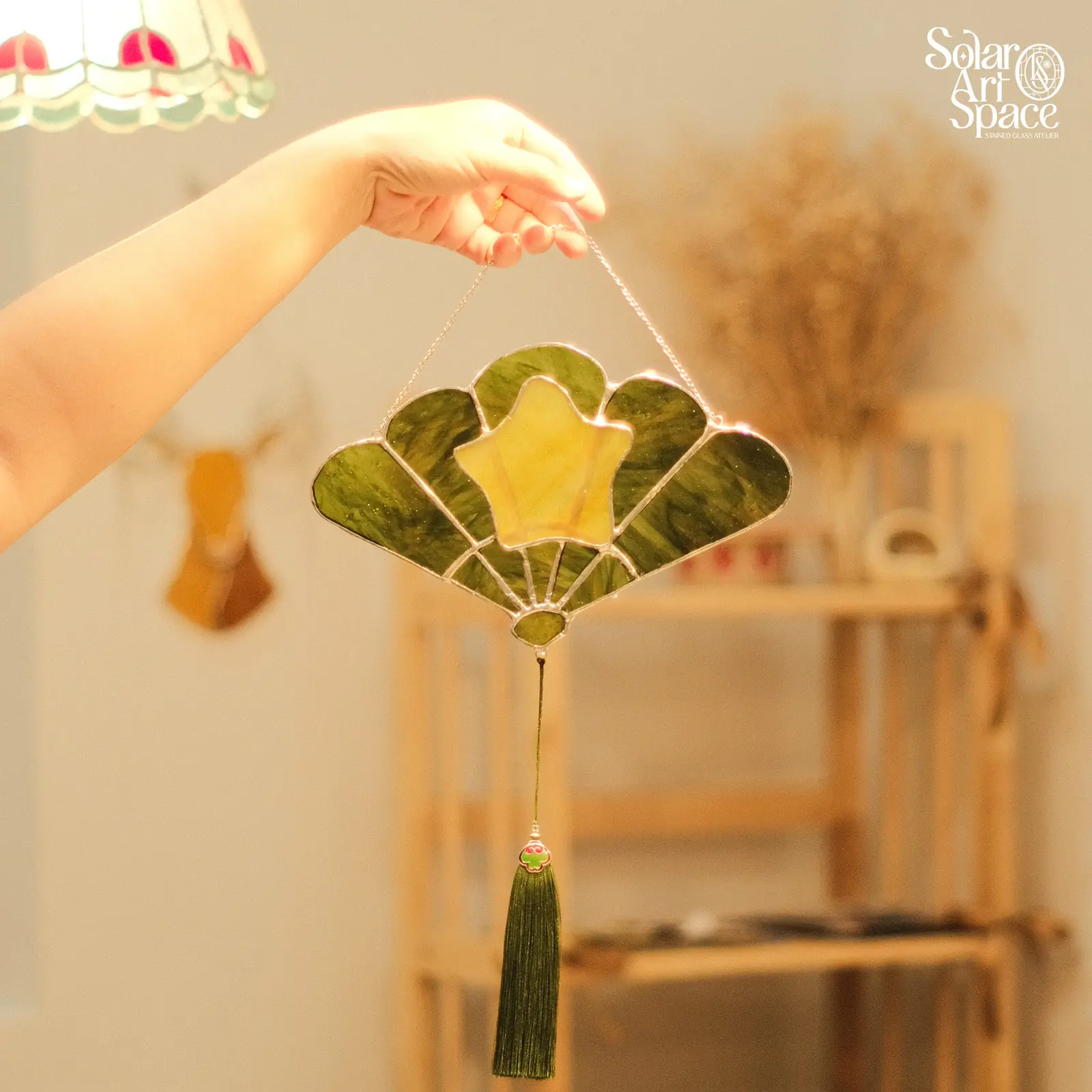 Suncatcher hình quạt xếp, kính xanh rêu có vân, ngôi sao vàng nhạt ở giữa, một sợi tua rua xanh buông xuống, treo trên dây xích
