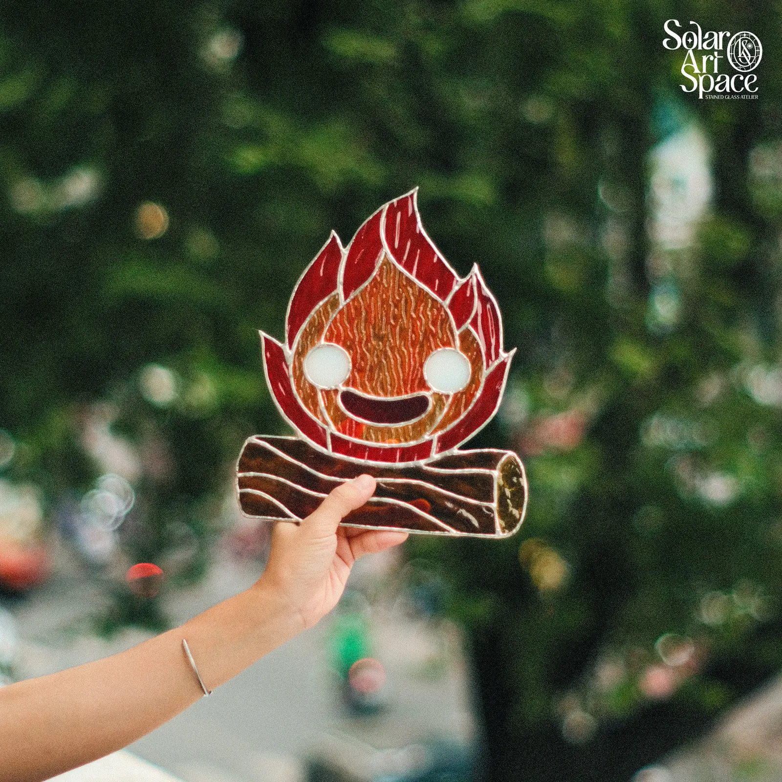 Suncatcher hình ngọn lửa đỏ cam có hai mắt và miệng cười, cháy trên hai khúc củi kính nâu, treo trên dây xích trước hàng cây