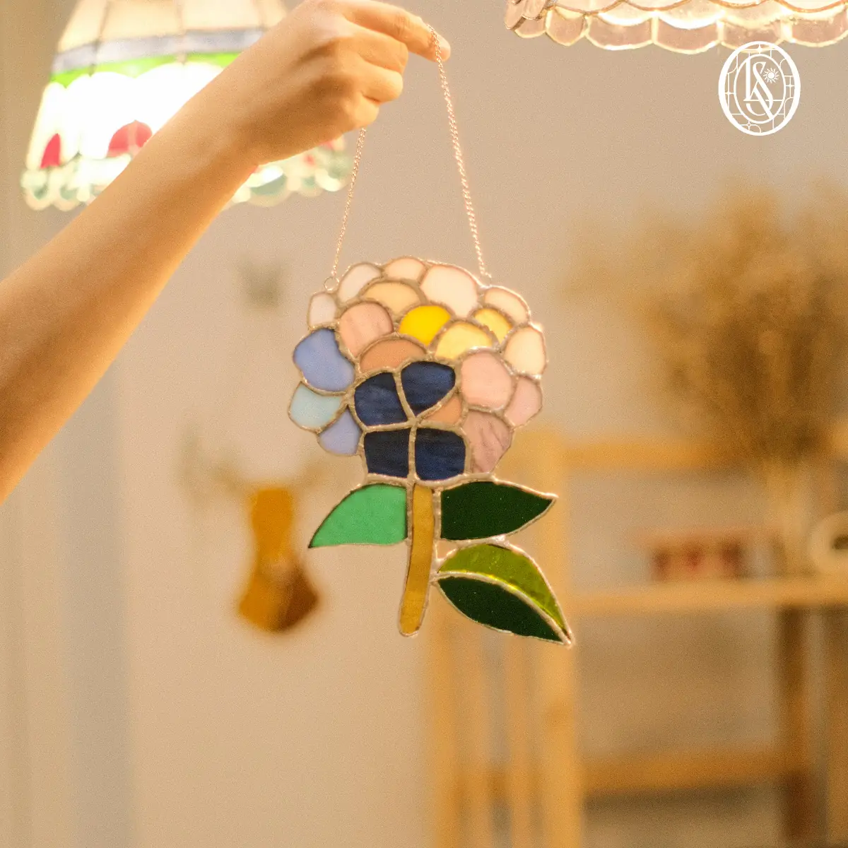 Suncatcher hình chùm hoa cẩm tú cầu, mỗi cánh một mảnh kính màu khác nhau — tím, hồng, vàng, xanh — ba chiếc lá xanh phía dưới, treo trên dây xích trong xưởng