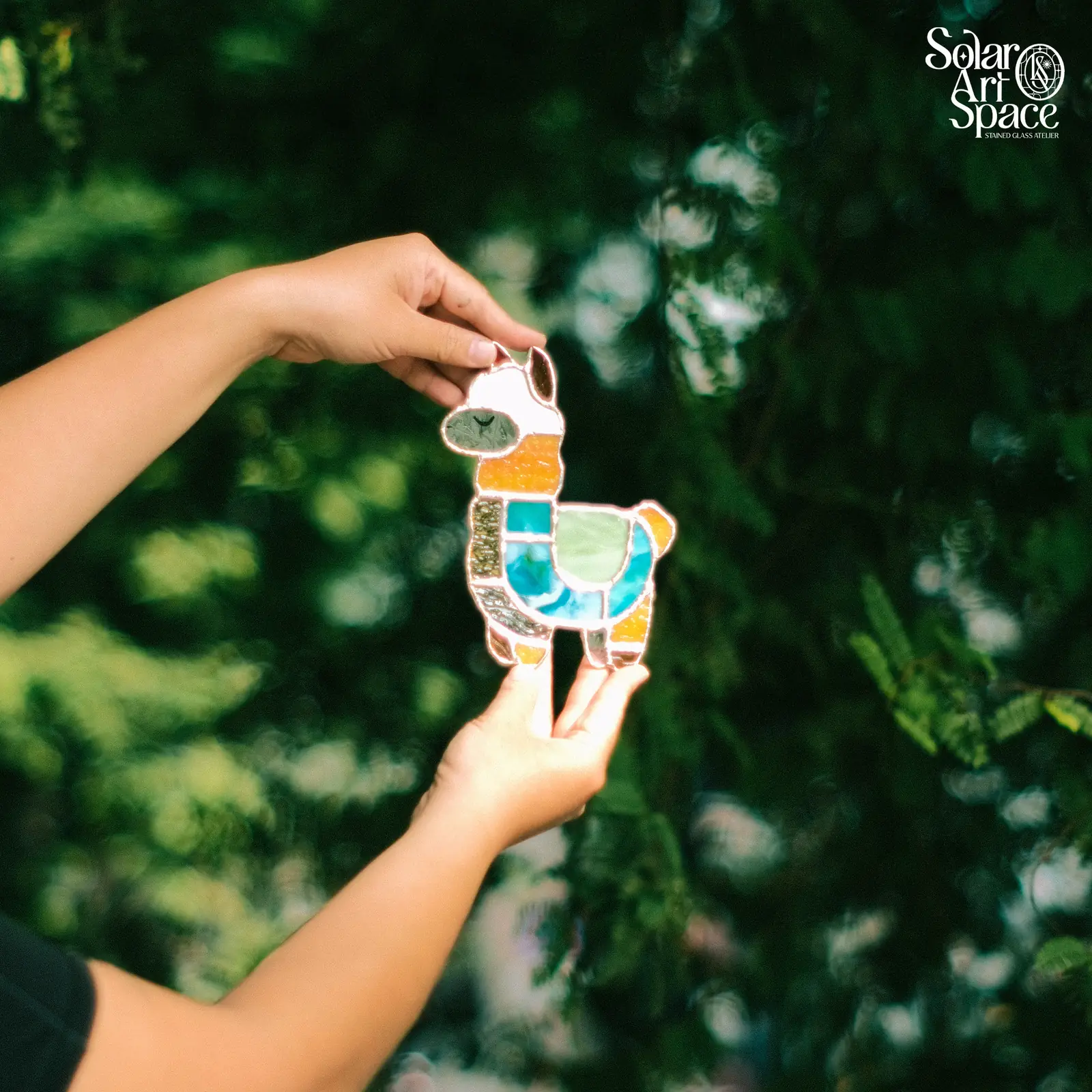 Suncatcher hình chú lạc đà alpaca đầu trắng, mõm xanh xám, cổ cam, thân ghép từ những mảnh kính xanh ngọc, xanh lá và vàng vân, hai tay cầm trước tán lá