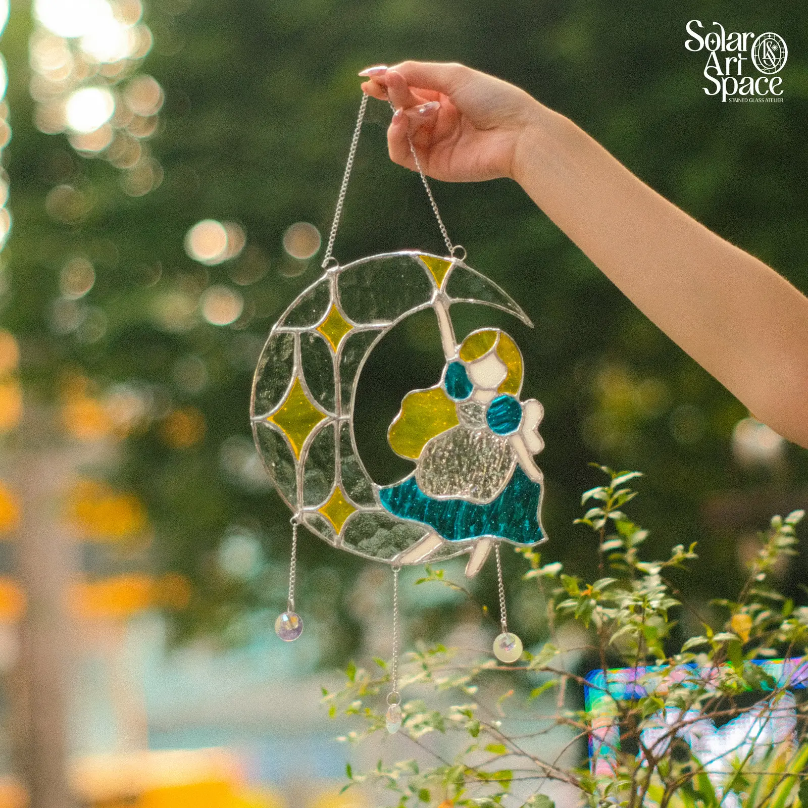 Suncatcher trăng khuyết bằng kính trắng có vân ghép nhiều mảnh, một cô gái tóc xanh ngồi ở đầu trăng, mấy ngôi sao vàng, hai viên đá rủ dưới bằng dây, cầm giơ trước hàng cây