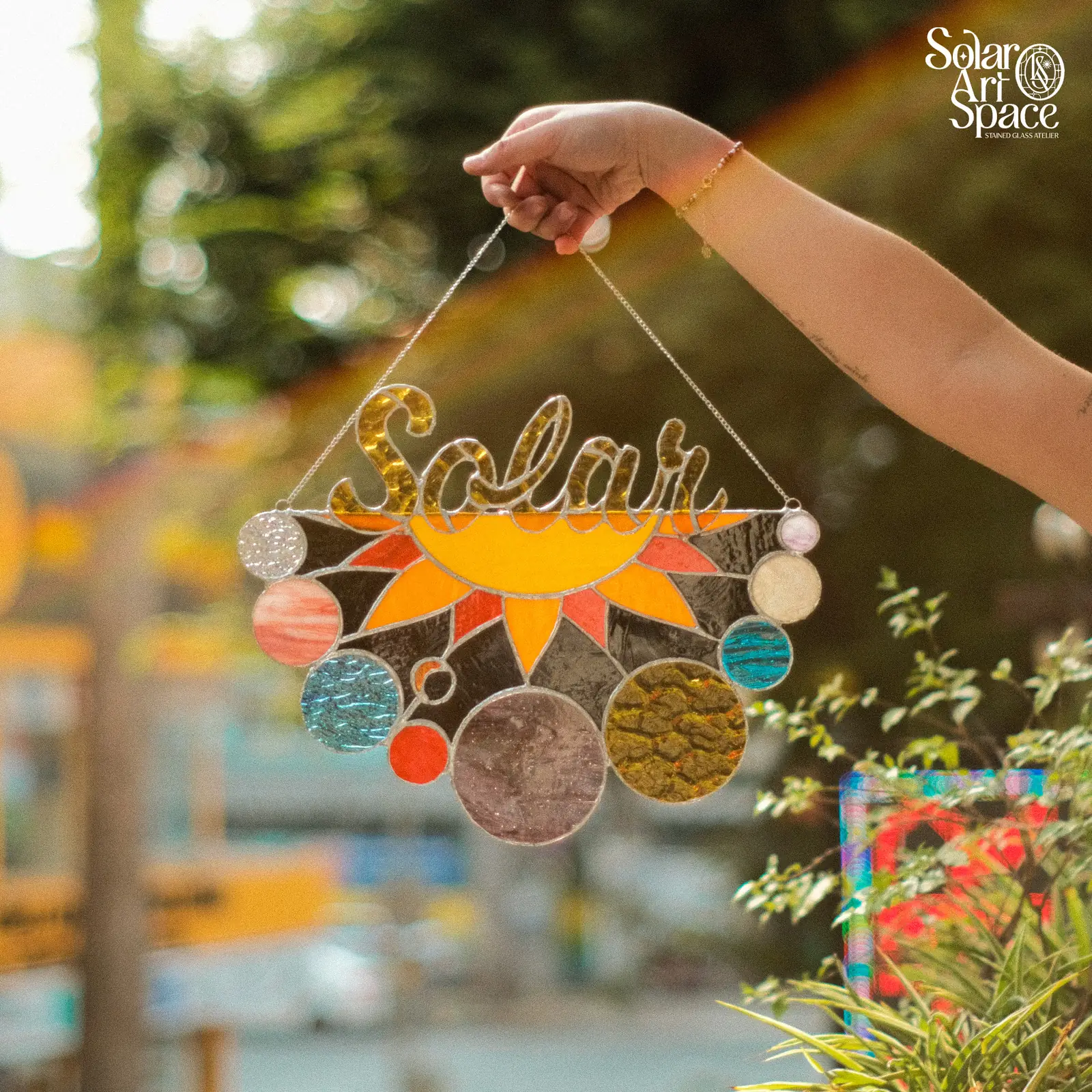 Suncatcher chữ Solar uốn bằng chì, bên dưới là mặt trời cam toả tia và một dãy hành tinh tròn bằng nhiều loại kính vân, cầm giơ trước hàng cây