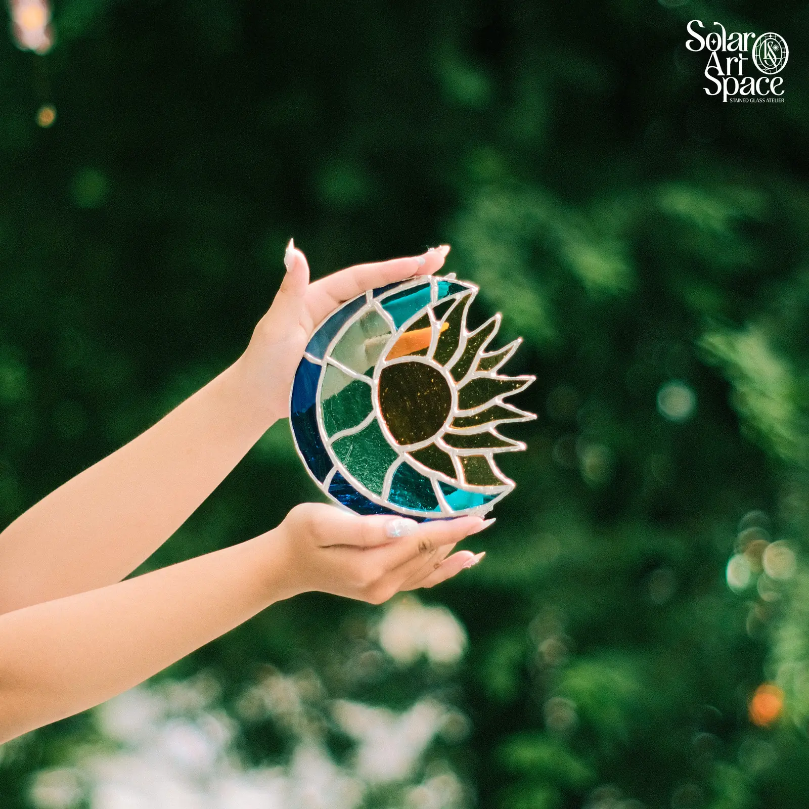 Suncatcher hình vầng trăng khuyết kính xanh lam ôm một mặt trời lõi kính nâu vân, tia nhọn xanh lá toả ra bên phải, hai bàn tay nâng trước tán lá
